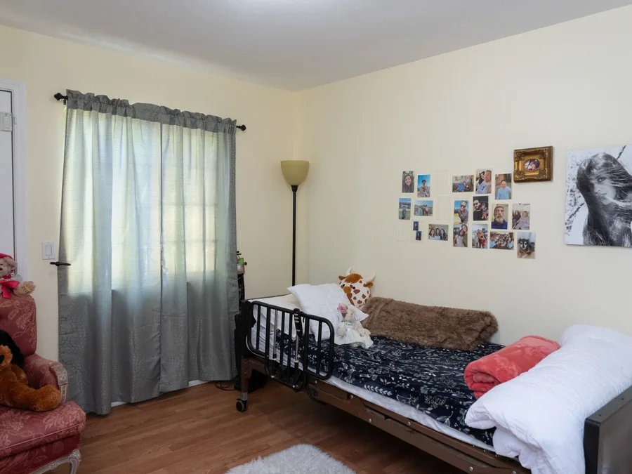 Comfortable resident bedroom with hospital bed, family photos on wall, and natural lighting from window with curtains
